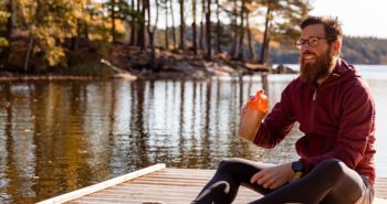 A man is sitting on an exercise mat outdoors by a lake. He is holding a protein shake.